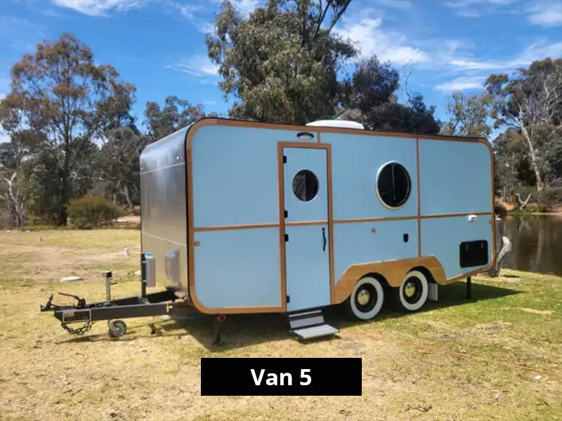Exterior of Van 5 tiny home cabin with light blue panels, timber trim, twin axle design, circular windows, and parked lakeside at Stawell C&C Park.