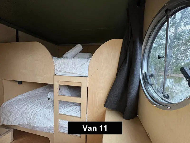 Compact camper van interior with wooden bunk beds, white bedding, and round porthole window, minimalist mobile accommodation design