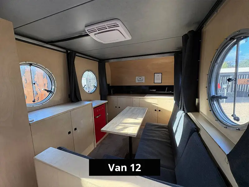 Retro van interior with dining area, built-in bench seating, wooden table, kitchenette, and round windows for natural light