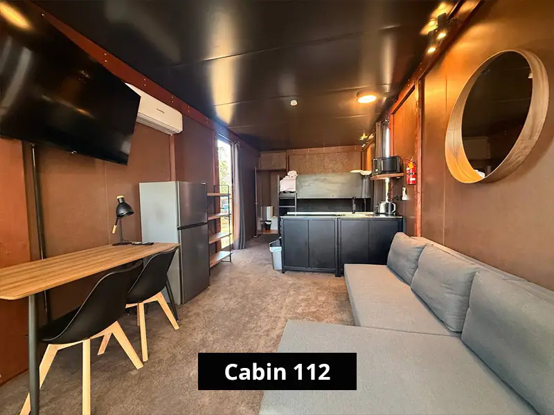 Cabin 112 bathroom with white toilet, vanity, black fixtures, large mirror, brown walls, and wood-look flooring.
