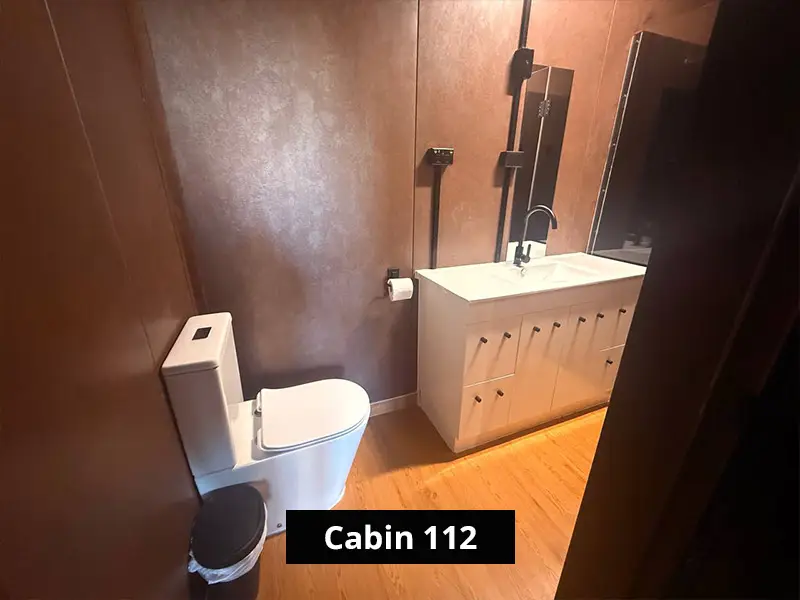 Private Cabin 112 outdoor deck with barbecue grill, metal chairs, black railing, and scenic riverfront bushland views.